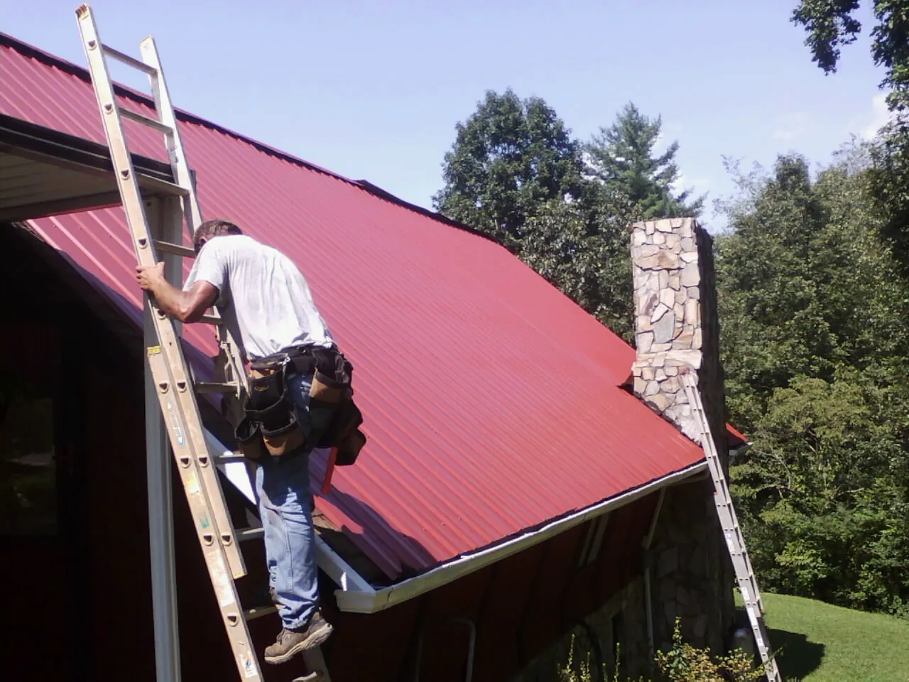 Roofer on a ladder beside a red metal roof and stone chimney — Soffit & Fascia Repair by Roofing Brothers in Oakes
